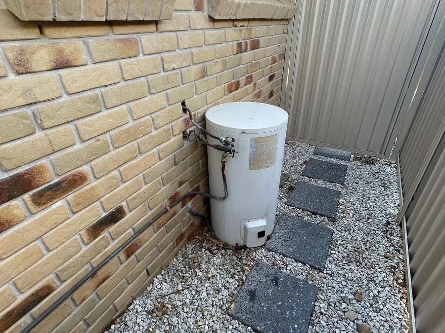 Small old electric hot water unit sitting on gravel beside a brick home