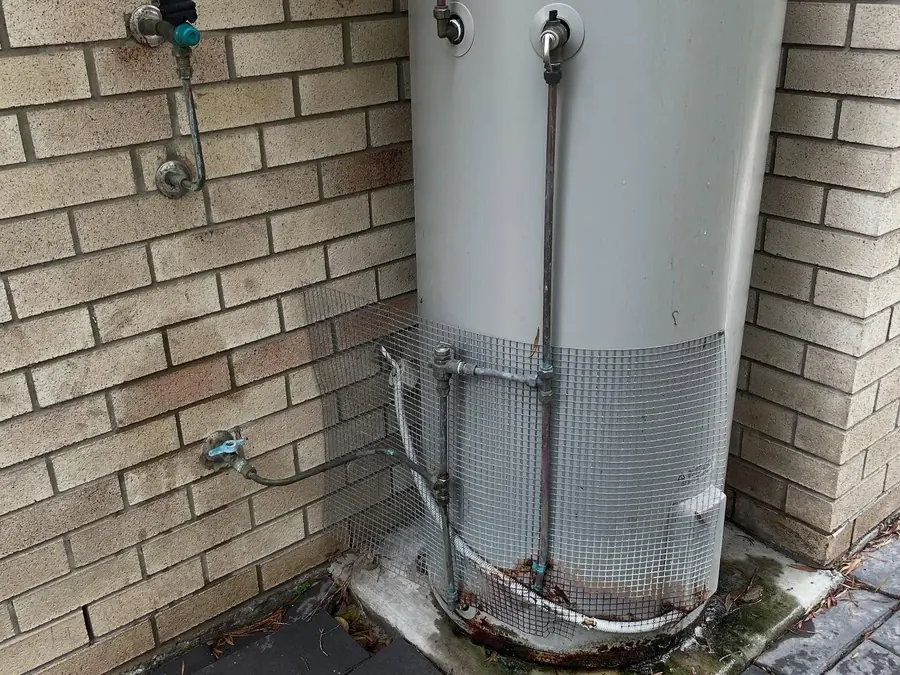 Old Vulcan electric hot water system with corroded pipework beside a brick wall
